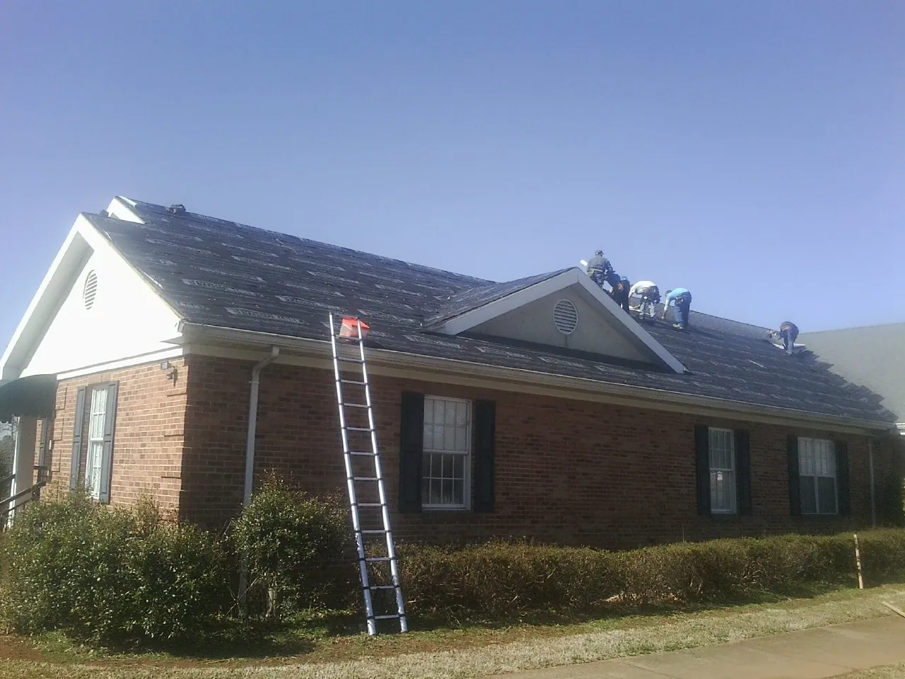 Crew tearing shingles off a brick ranch roof with a ladder set — Emergency Roof Repair work by our crew in Woonsocket