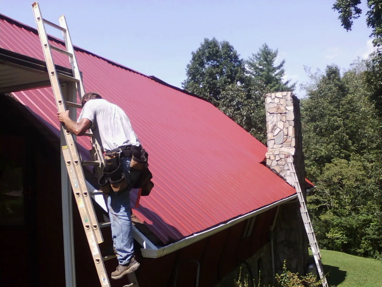 Roofer on a ladder beside a red metal roof and stone chimney — Soffit & Fascia Repair by Roofing Brothers in Woonsocket
