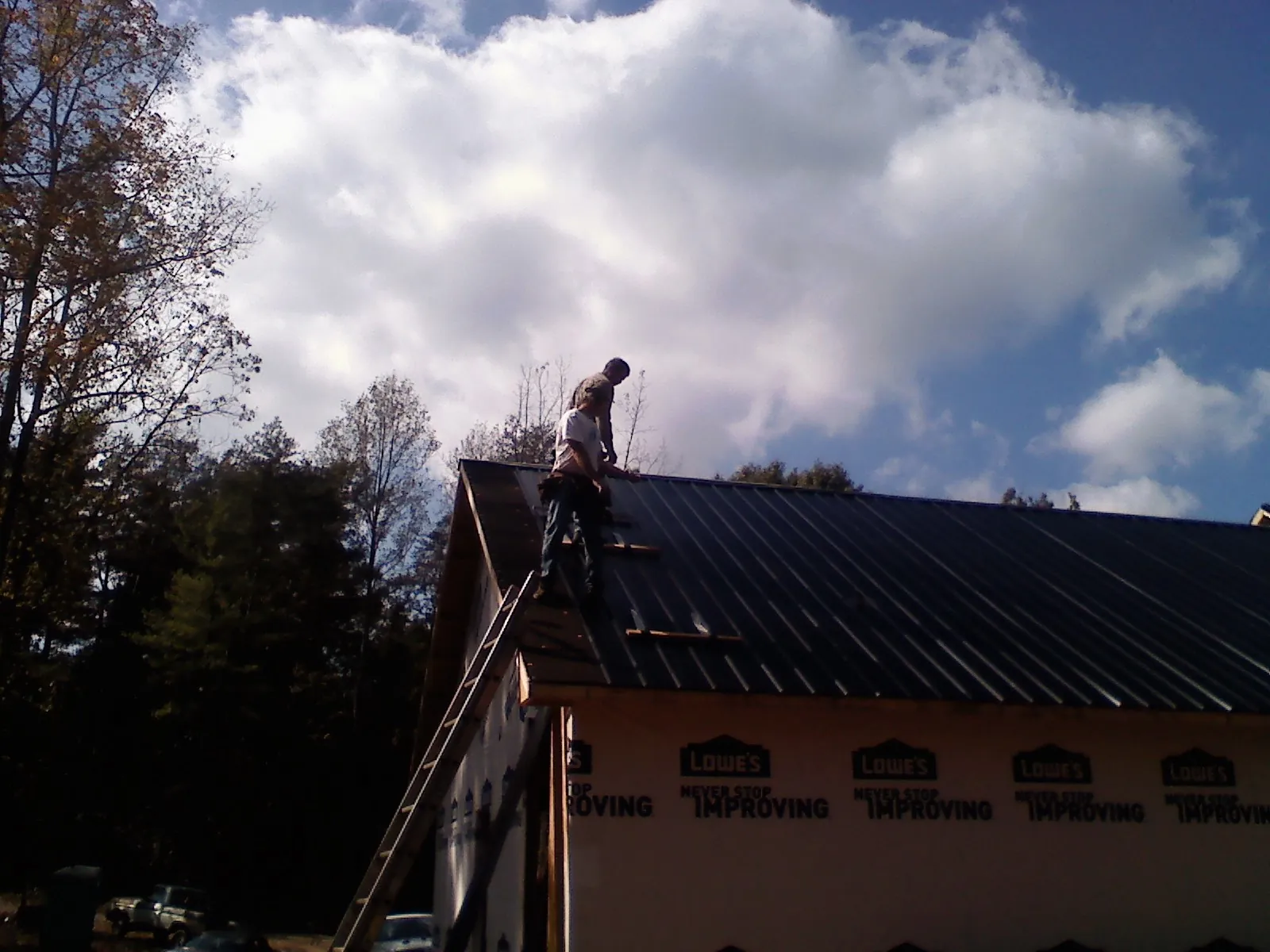 Roofer fastening dark metal panels high against a cloudy sky, from a Roof Inspection job in Woonsocket
